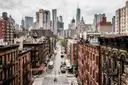 A downtown Manhattan street of brick tenements seen from above, with Lower Manhattan towers behind