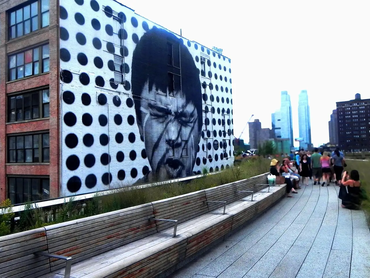 Visitors on the High Line's wooden path next to a large mural, with Manhattan towers behind