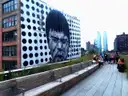 Visitors on the High Line's wooden path next to a large mural, with Manhattan towers behind