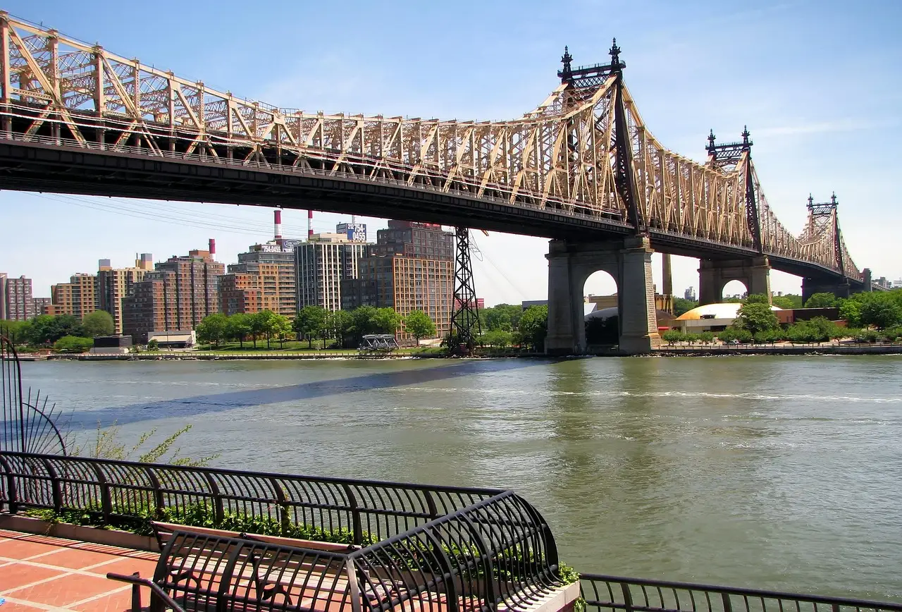 The Queensboro Bridge crossing the East River towards Queens on a sunny day, seen from a riverside promenade