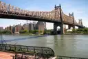 The Queensboro Bridge crossing the East River towards Queens on a sunny day, seen from a riverside promenade