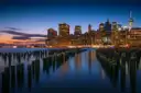 Lower Manhattan at dusk seen past old pier posts on the Brooklyn shore
