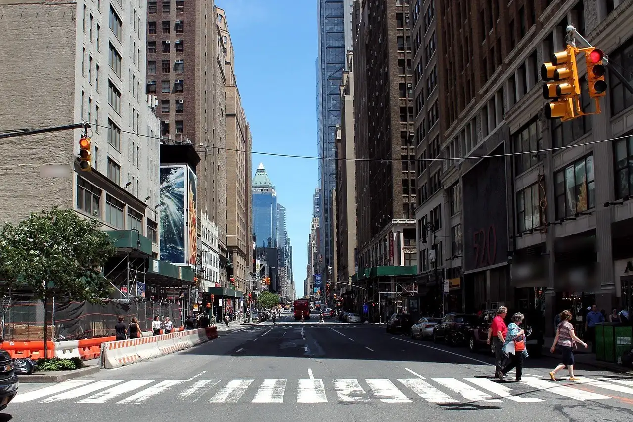 People walking over a wide striped crosswalk on a long, straight Manhattan avenue on a sunny day